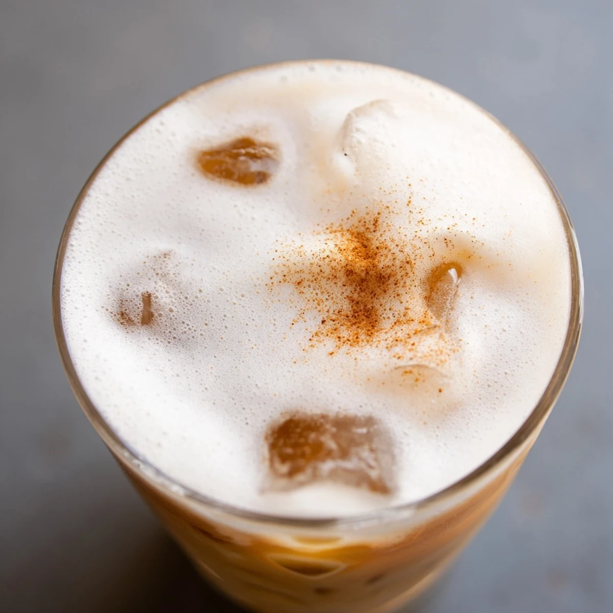 Chilled iced chai tea with cold foam layered over ice cubes, garnished with cinnamon in a clear glass on a sunny tabletop.  