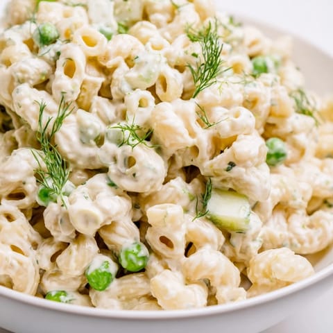 Chilled dill pickle pasta salad with a creamy dressing, served in a white bowl and garnished with fresh herbs.
